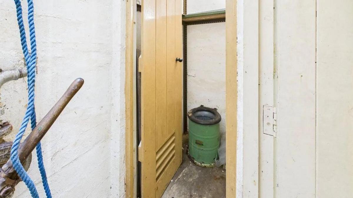 Interior of the bunker, showing the toilet. The walls are painted white and the floor is made of concrete. A yellow painted door is propped open to show a green metal chemical toilet. In the foreground, is a wooden handle and a blue nylon rope. To the right is another white painted door.