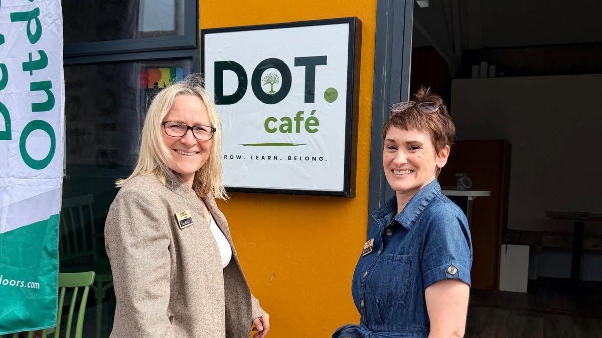 Two women stood in front of an orange building with a sign behind them reading "DOT. cafe".