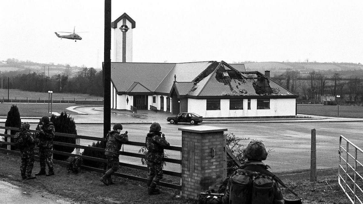 A helicopter is flying in the sky close to a damaged church. Soldiers are behind a fence. 