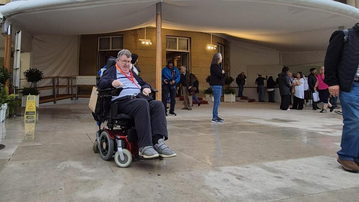 A man with grey hair and glasses sat in a wheelchair outside underneath a large tent