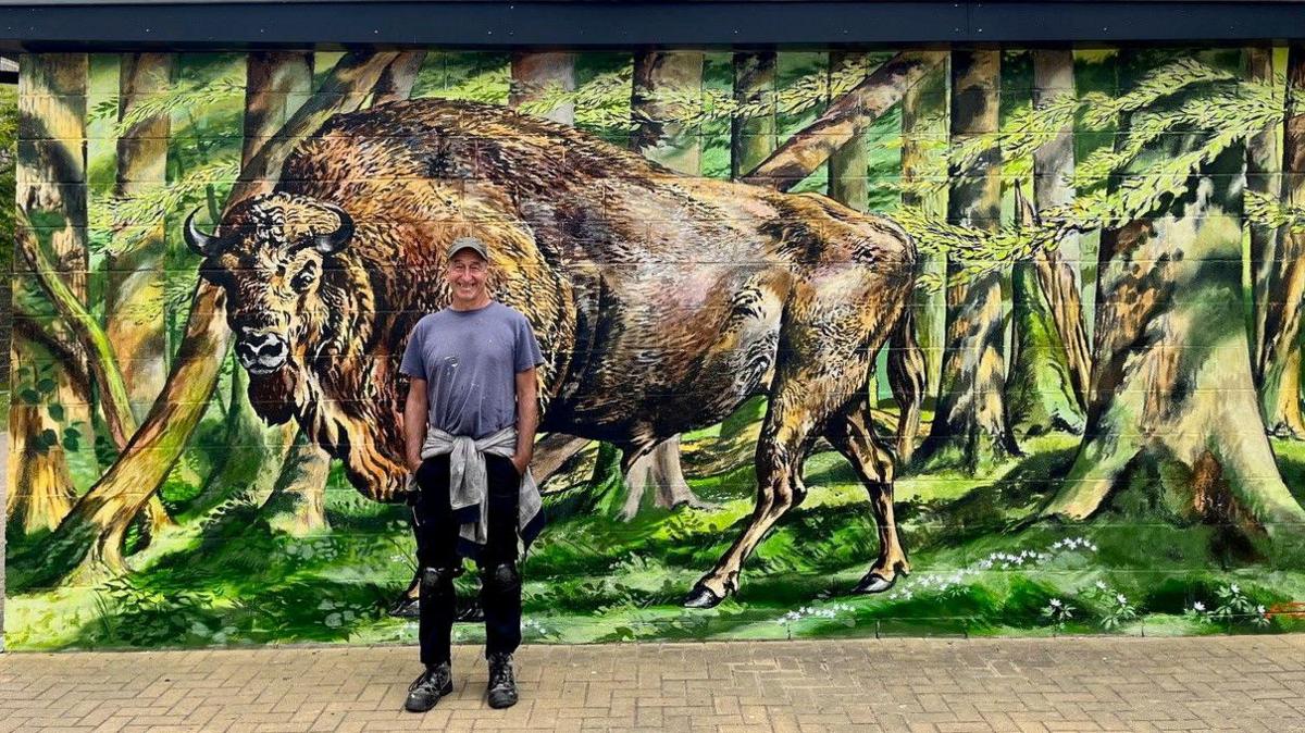 Street artist Mark Anthony standing in front of a mural of a bison outside the Gulbenkian Theatre at the University of Kent.