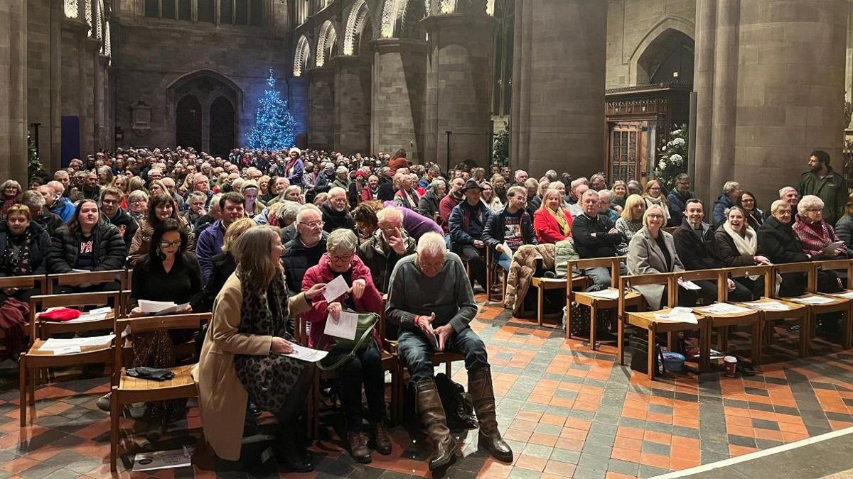 A large number of people inside Hereford Cathedral sitting on wooden chairs with the grey stone walls of the cathedral around them including huge stone pillars. The floor is tiled with red and black tiles in a diamond pattern.