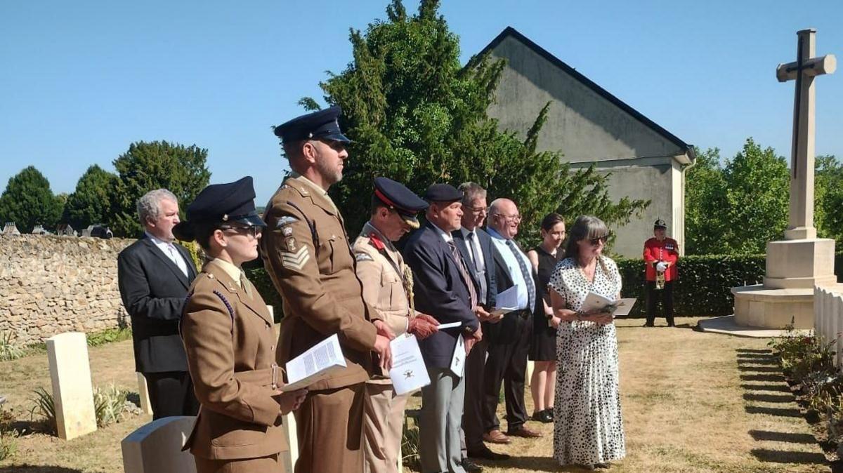 A group of people stand looking at a line of white graves. A few of the people are dressed in service uniform and the others are in suits and dresses. It is a sunny day and all their faces have solemn expressions.