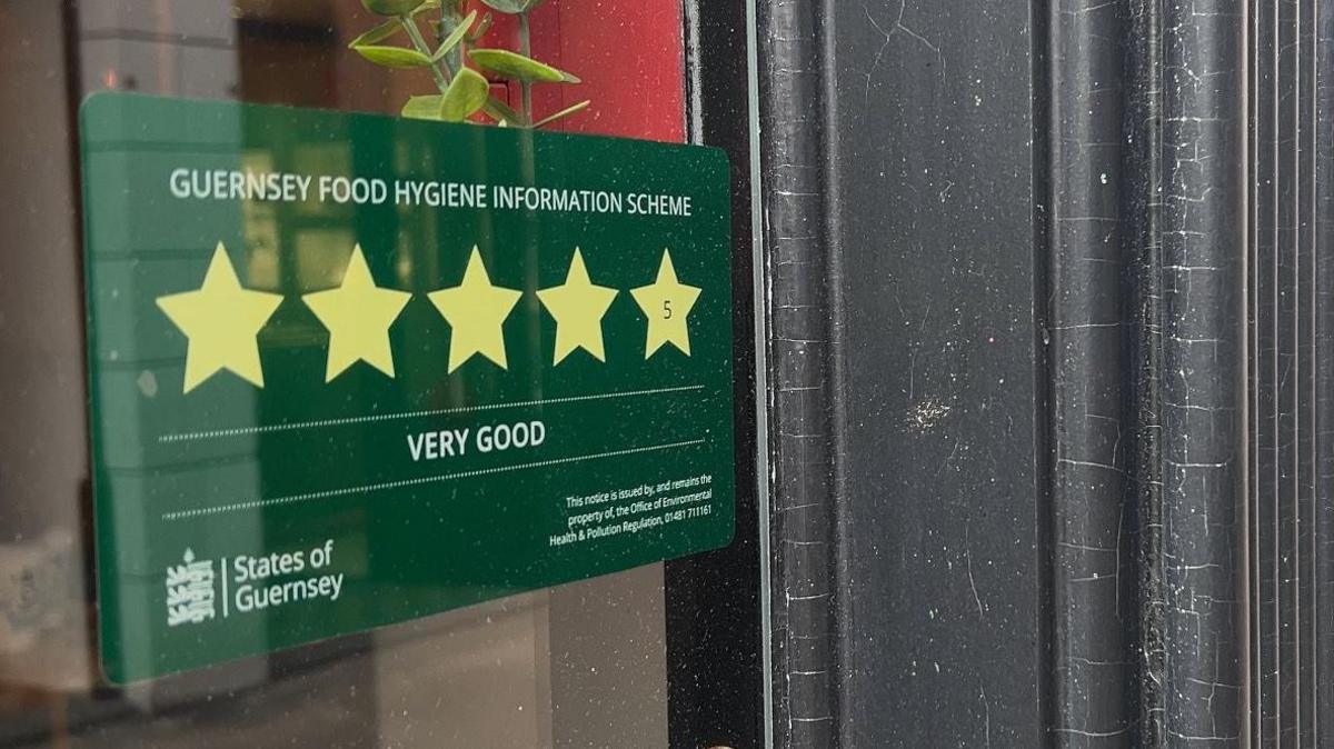 A photo of a green sticker on a window. The sticker has five stars on it and is titled 'Guernsey Food Hygiene Information Scheme'