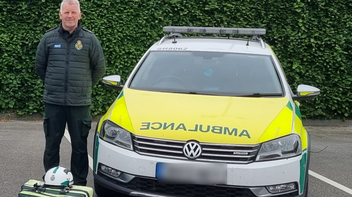 A man in a paramedic's uniform stands in a car park next to a car which says ambulance on it