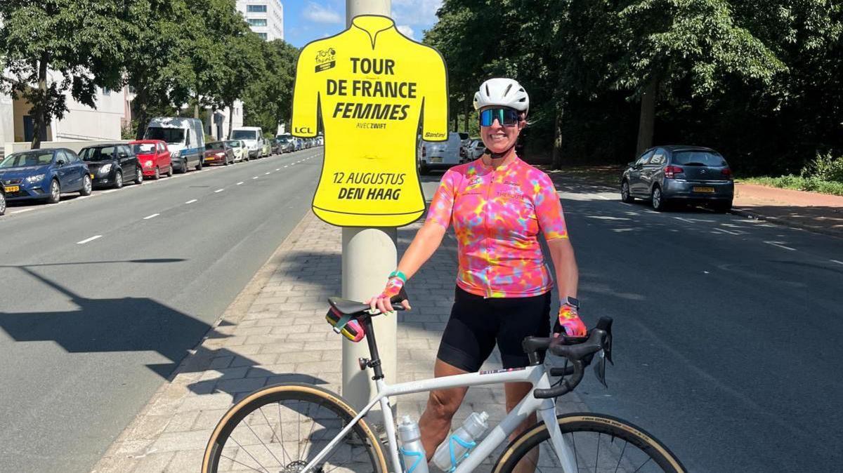 A woman wearing a pink short sleeved cycling shirt with matching gloves, black cycling shorts, big sunglasses and a white helmet, smiling holding her white bike next to a sign shaped in a yellow shirt saying 'TOUR DE FRANCE FEMMES', background of two roads with cars parked along the side, cloudy blue skies