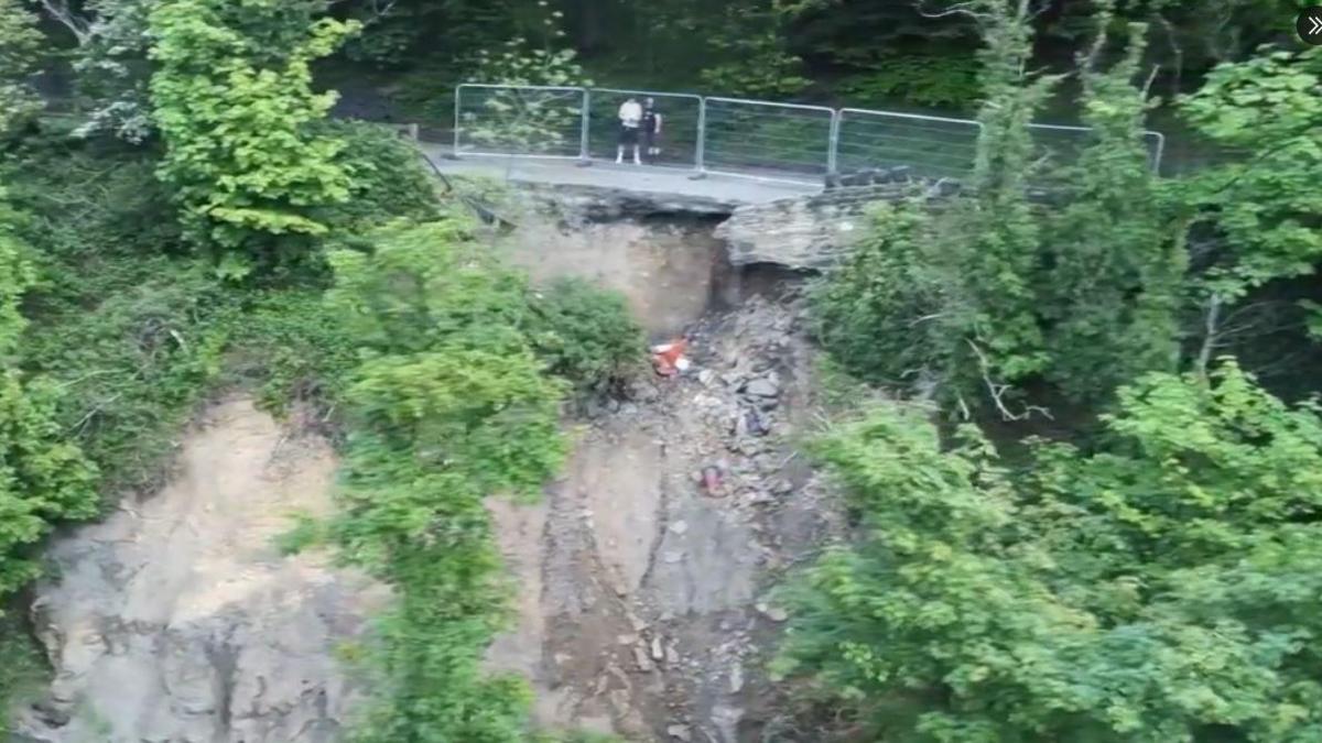 Two people standing on the road near a section which has eroded and fallen into the river below