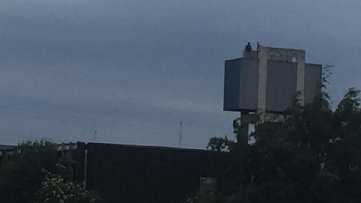 A blurry photograph of a rectangular, concrete water tower on grey stilts with two human figures standing on the top. There is tree foliage and metal fencing in the foreground.  