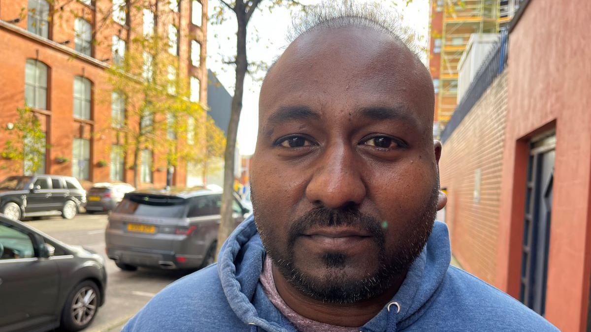 Delivery driver Ramakrishnan Kannan pictured wearing a blue zip up jumper. It is a close up shot but you can see cars parked on the side of the street in the background.