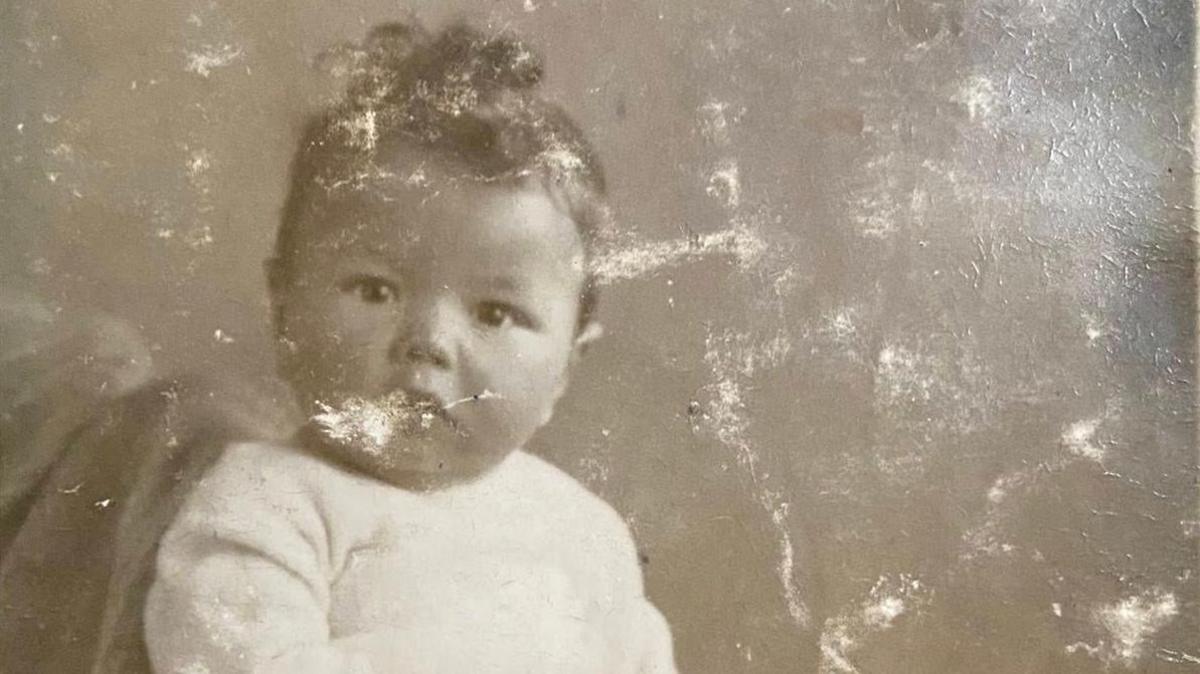 An old sepia photograph of a young child. The boy looks directly at the camera and has dark curly hair and large brown eyes. He wears a white jumper and the picture is cracked white and appears weathered.