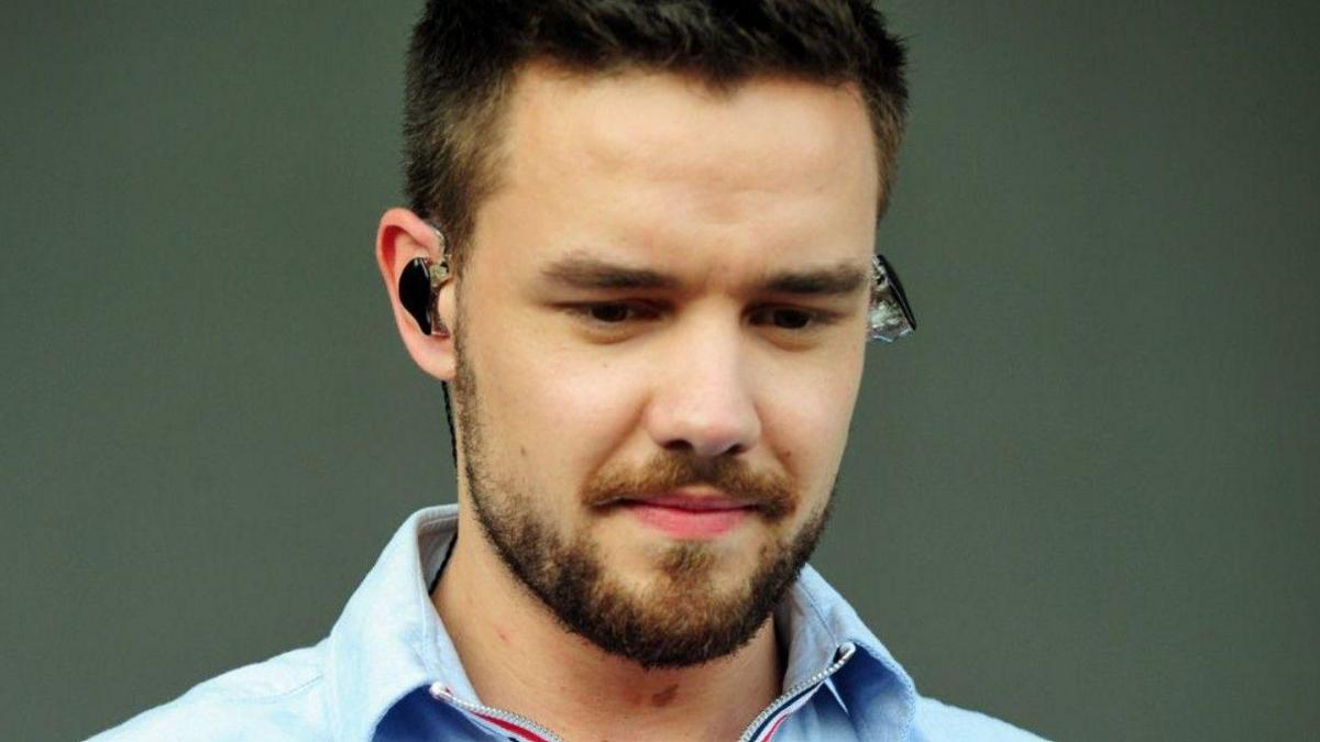 Liam Payne, who has short dark hair and a beard, is wearing an earpiece and a blue shirt and is looking down. He is standing against a grey background.