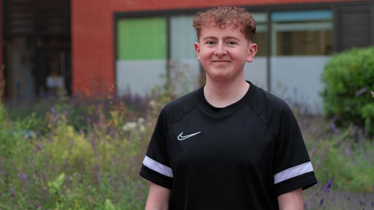 Harrison Sherwell, with short brown curly hair is wearing a black casual fit t-shirt with a thick white horizontal stripe on the sleeves. He is smiling, standing against a backdrop of wildflowers.