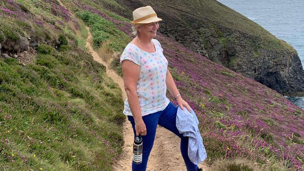 Jo Kehoe Butler, wearing a white top, blue jeans and a hat, standing on a coastal cliff path. There are purple flowers around her and the sea is in the distance.