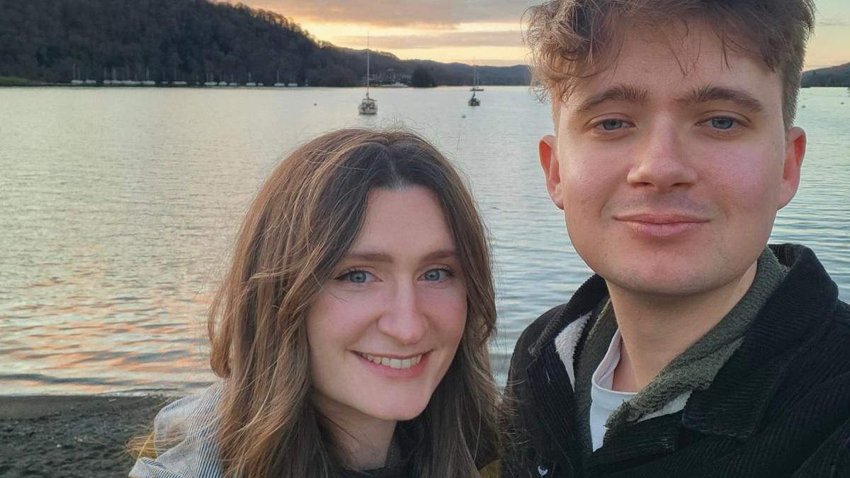 Emily, left, and Sam, right, look at the camera as they take a selfie in front of a lake at sunset. A couple of small boats can be seen bobbing on the water in the background.