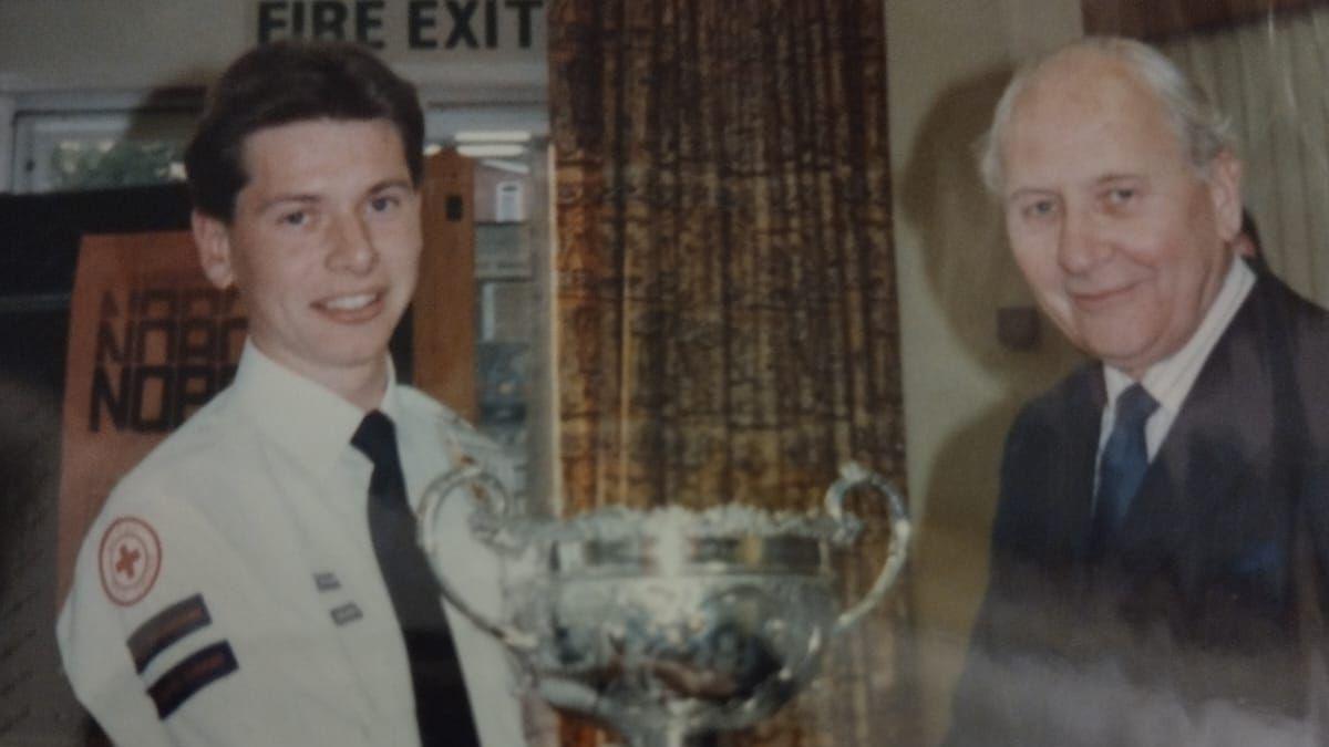 Man in Red Cross uniform with a silver trophy stands with a man in a navy suit.