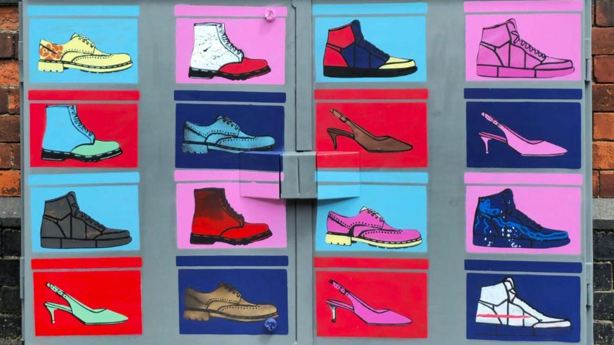 A colourful piece of artwork on a utility box, painted with shoes and boots. The main colours are purple, pink, blue and red. They are 16 boots and shoes, all in four rows, with four images in each row. A brick wall is behind the box.