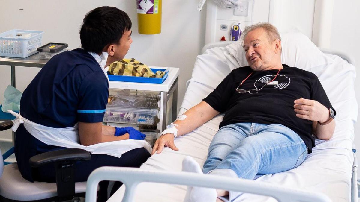 A hospital room with a male patient lying on a bed and a healthcare worker in scrubs attending to the patient's arm. The patient is dressed in blue jeans and a black t-shirt, and appears to be receiving medical attention. Medical equipment, including a trolley with medication and supplies, are visible beside the bed.