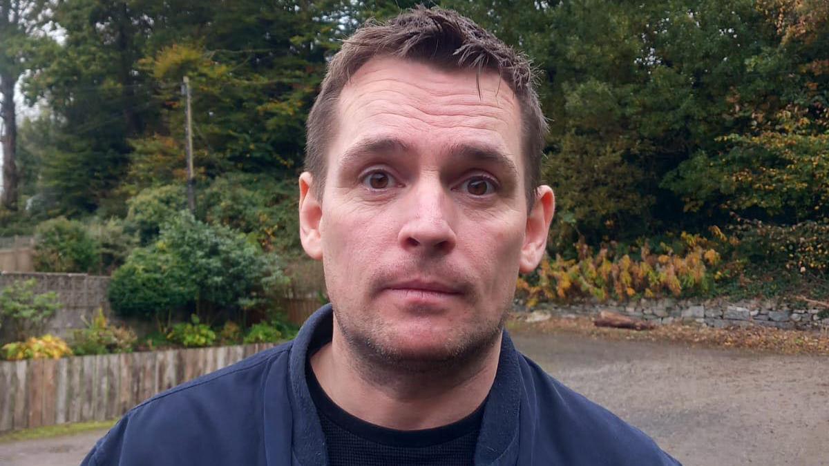 A man with brown, spikey-fringed hair and brown eyes in a black T-Shirt and blue jacket stands in front of a driveway with plants and a fence in the background