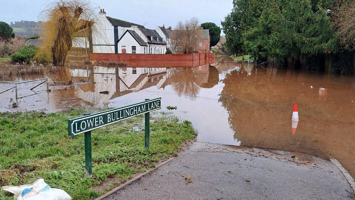 A road sign reading Lower Bullingham Lane, with pavement and grass area flooded with brown water. A house in the background is surrounded by a red brick wall, but the flooding extends past the house.
