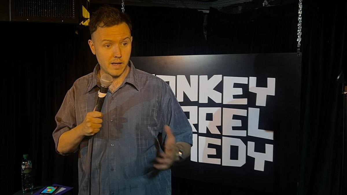 A comedian stands on a darkened stage with obscured white lettering on the back wall.