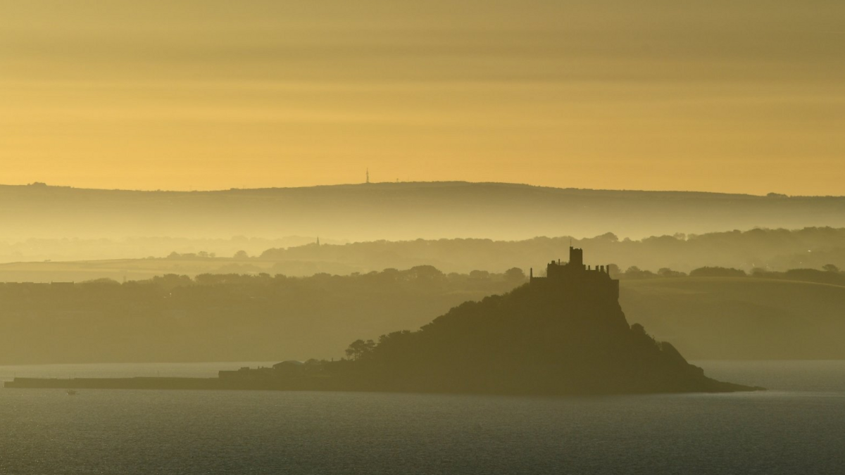 St Michael's Mount is seen shortly after sunrise. The sky is yellow/orange and the mount is silhouetted against the backdrop.