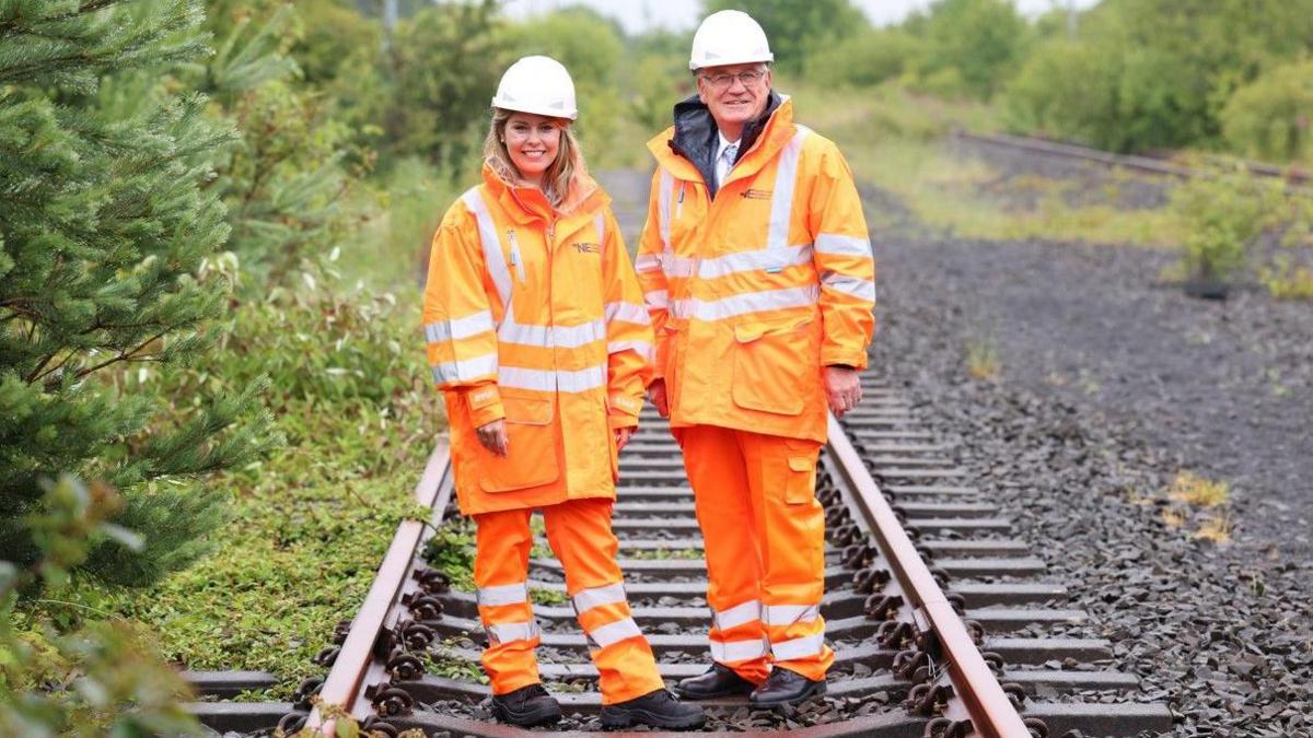 A woman with blonde hair and a man with glasses are dressed in orange Hi-Viz clothing, white hats and standing on a disused railway track