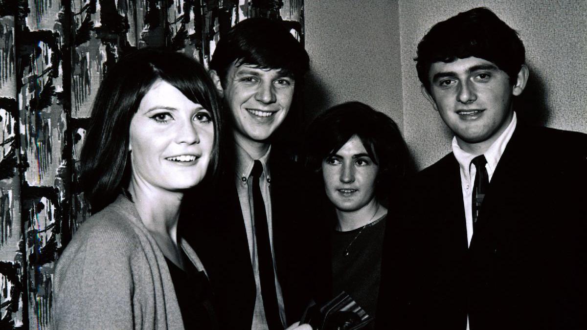 Black and white photo of sanide shaw signing autographs. in the background are two men in 60s style suits and a woman wearing a dark dress