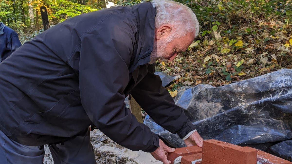 A man, with grey hair and a grey beard, placing bricks on a wall. He is bending over, has a dark coat on and dark trousers. Foliage, leaves and plastic sheeting is behind him. 