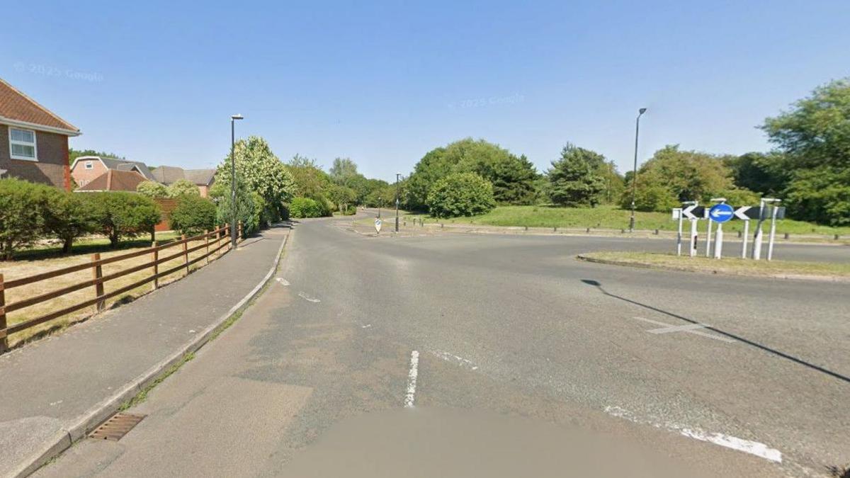 Small roundabout with several roads leading off it and one dark-coloured car driving off the roundabout to the right