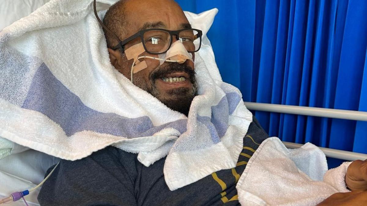 Earl Falconer lies in a hospital bed eight days after coming off a ventilator. He is wearing glasses and has a nasal tube attached, with a towel surrounding his head