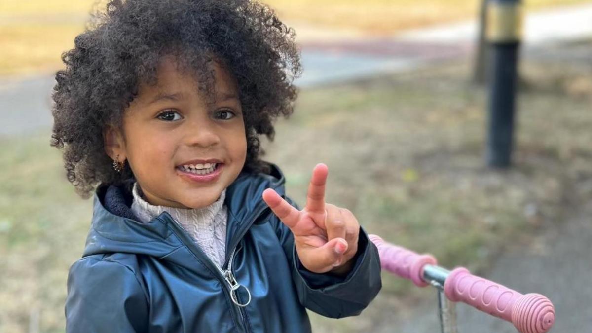 A young girl with brown curly hair doing a peace sign while standing and smiling in a park