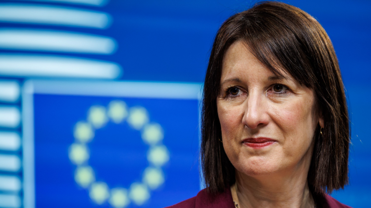 UK Chancellor Rachel Reeves stands in front of a blue and white EU flag She wears a burgundy suit and her fringe is swept to the side. 