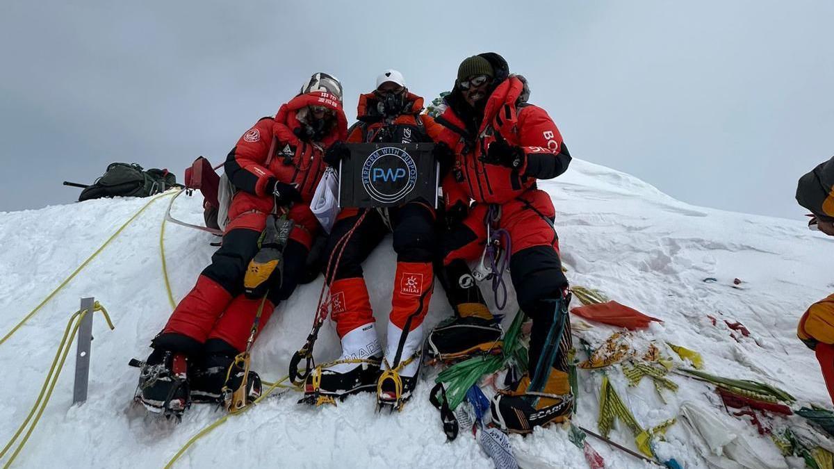 Three men in mountain gear standing at the top of a snowy slope with a grey sky behind them