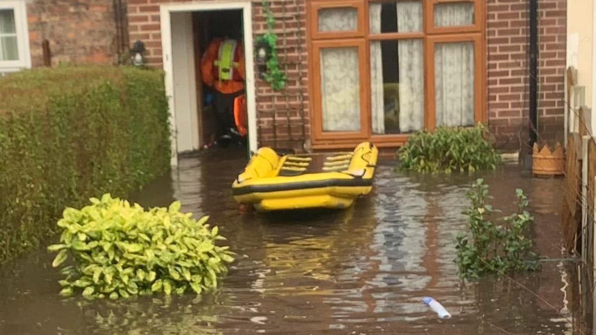 A yellow inflatable raft floats on brown muddy floodwater in the submerged back garden of a house