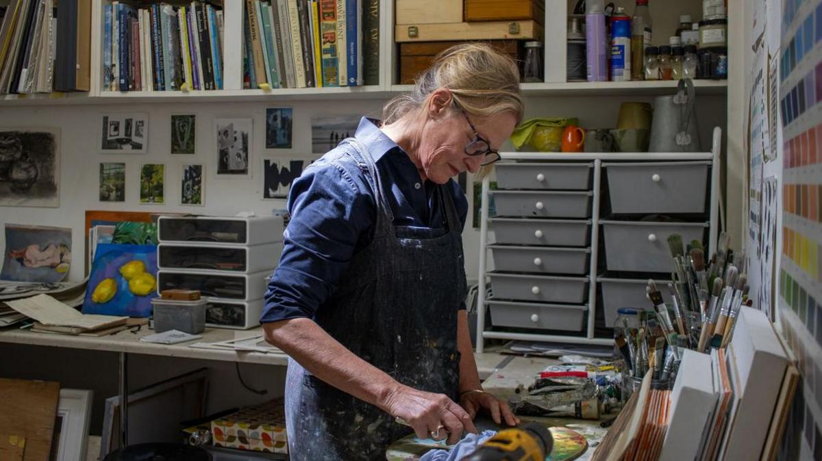 Woman working in blue overalls, standing with paintbrush in front of a canvas. There are colour charts on the wall and lots of drawers in the background.
