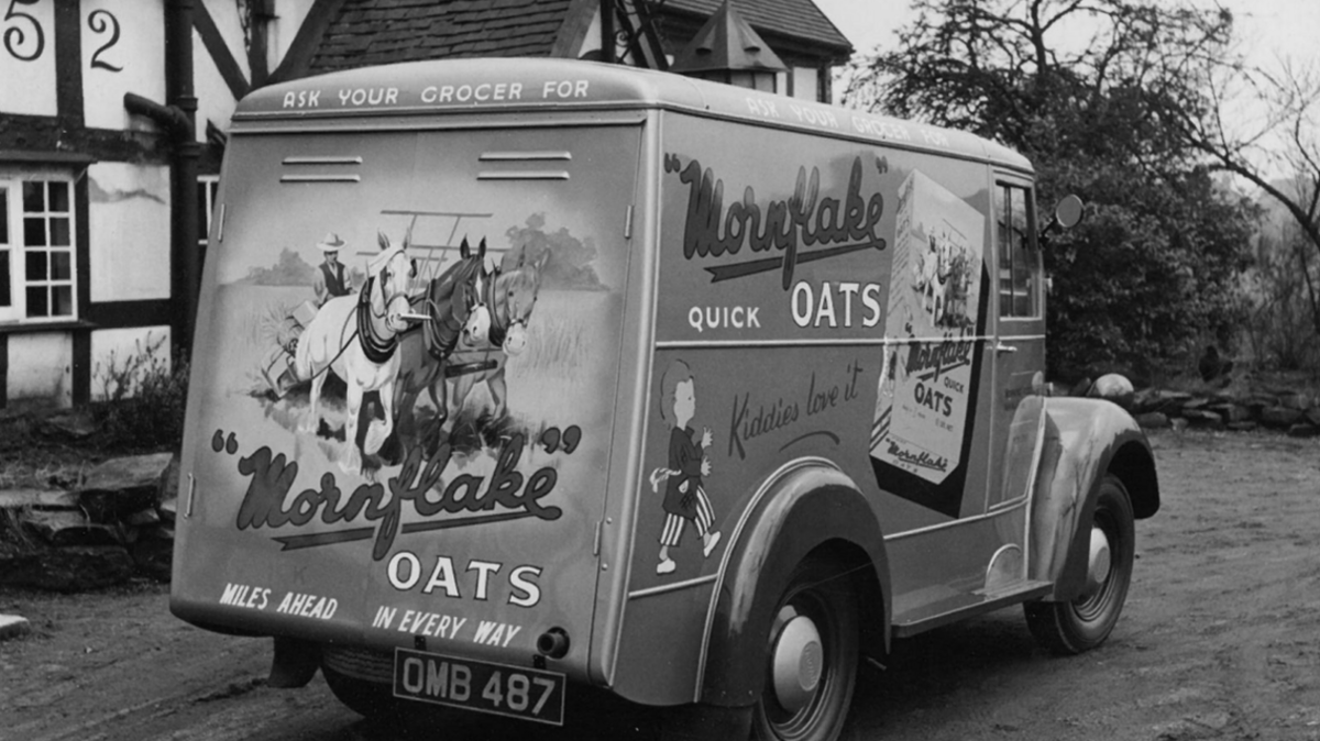 A black and white image showing an old-fashioned van with Mornflake branding on the side. It is parked outside a timber-framed house.