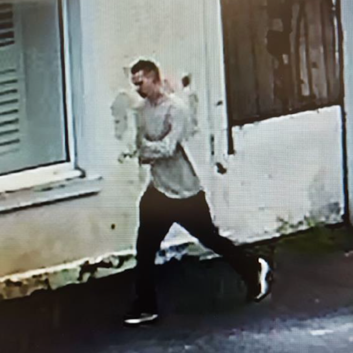 Aleksandr is walking along a street. He is wearing trainers, black jeans and a beige top. 