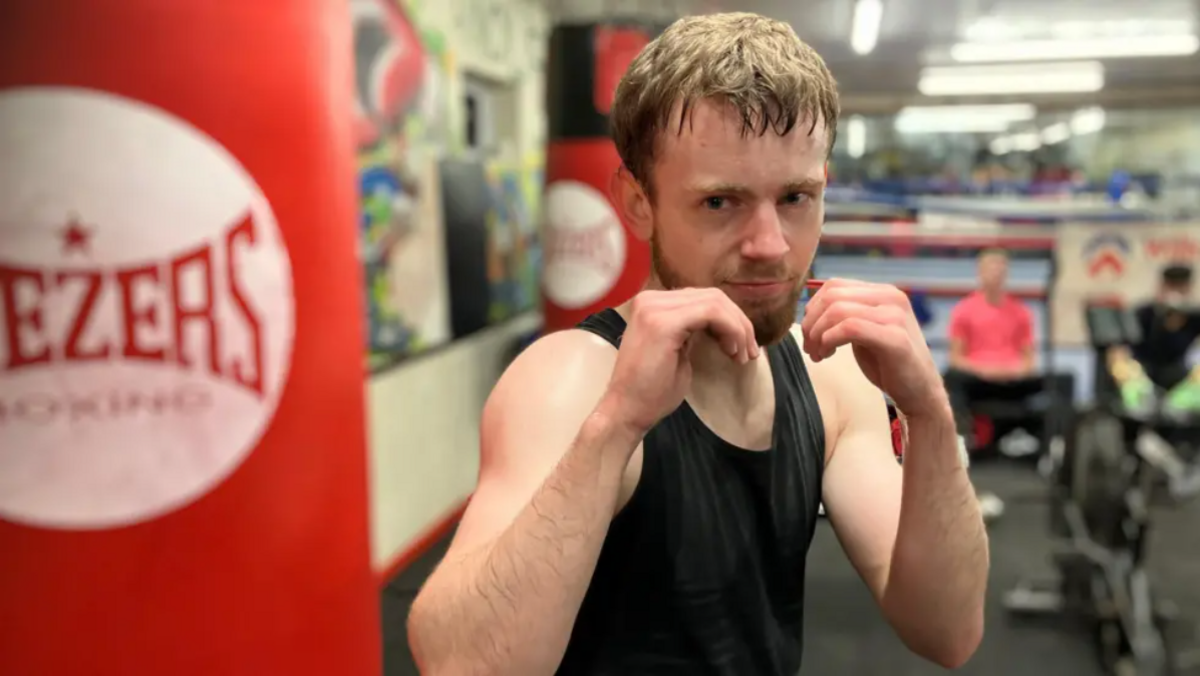 A young white man in a black, sleeveless top strikes a boxing pose, with hands up, in front of a red punching bag. He is pictured in a gym.