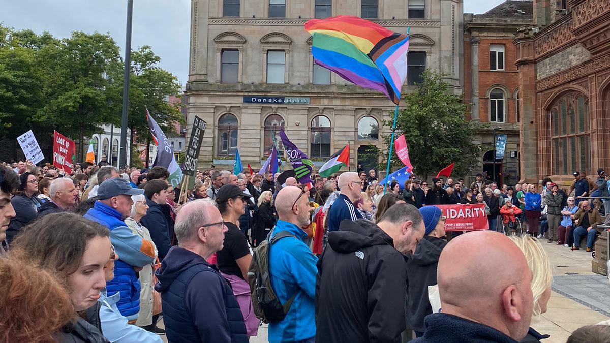 Flags are waved and people face the Guilldhall looking pensive, some wave flags and some stare ahead. Faces fill the picture, mostly middle aged men but some young women and flags vary in colour and size. 