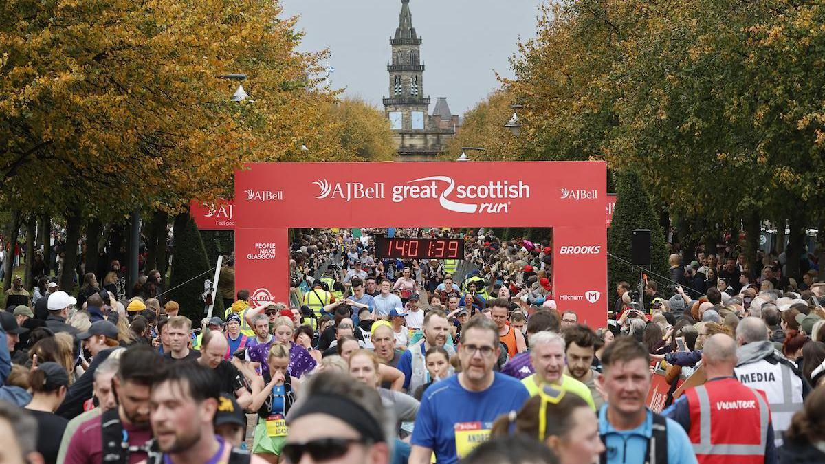 The AJ Bell Great Scottish Run, showing a large group of runners crossing the finish line under a red banner. The digital clock reads 1:40:13.9, and the scene features autumn trees and a historic building with a clock tower in the background.