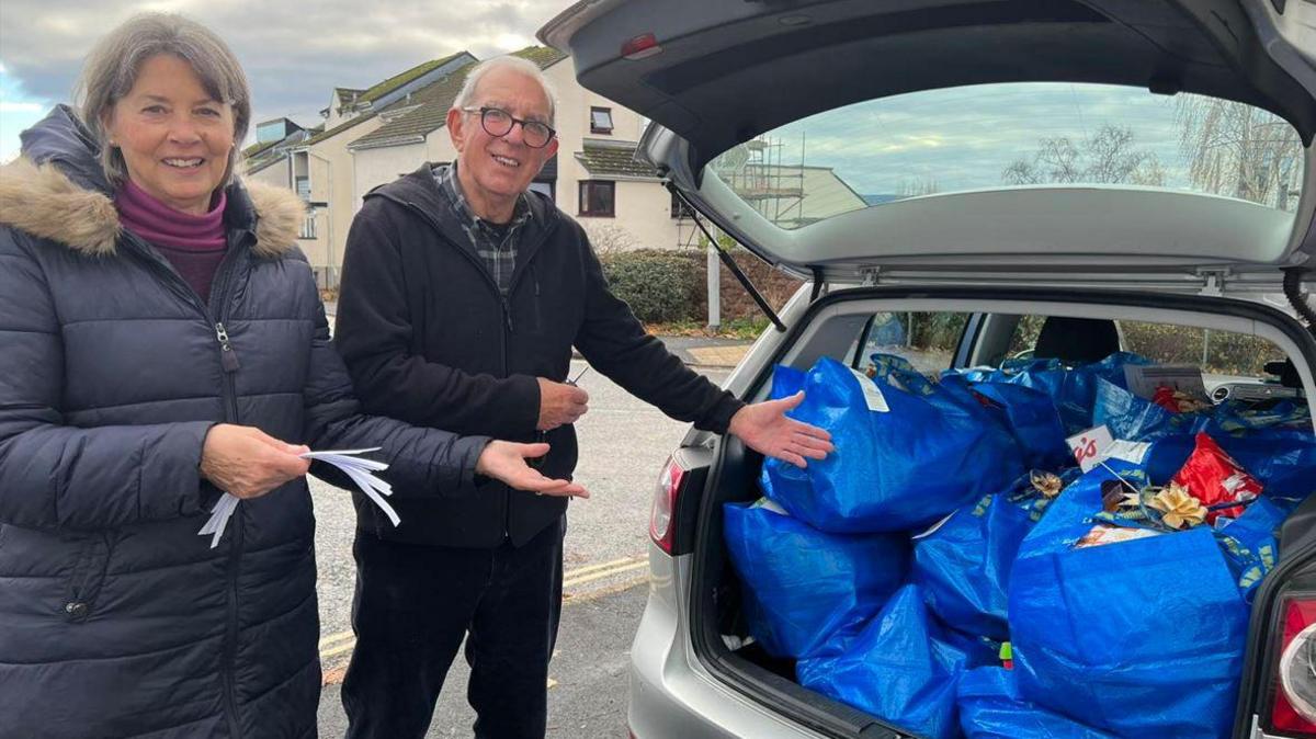 A women and men signallling to their car book which is full of Christmas hampers