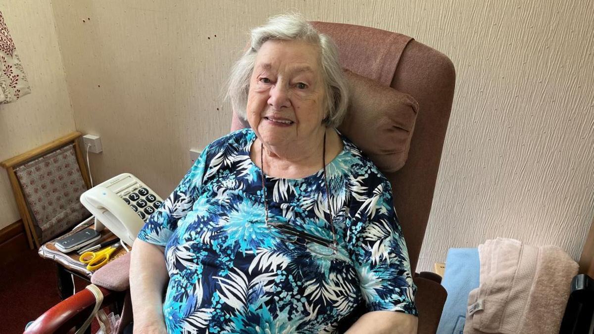 Dorothy Heron sitting in a chair in her home. She is wearing a blue top with a pattern of leaves. She has short white hair and is smiling