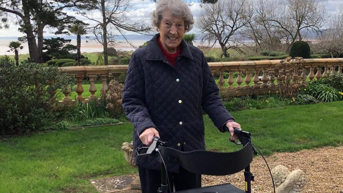 Joy Palfreman is standing in a garden with a walker. She is wearing a navy blue jacket and is smiling at the camera.