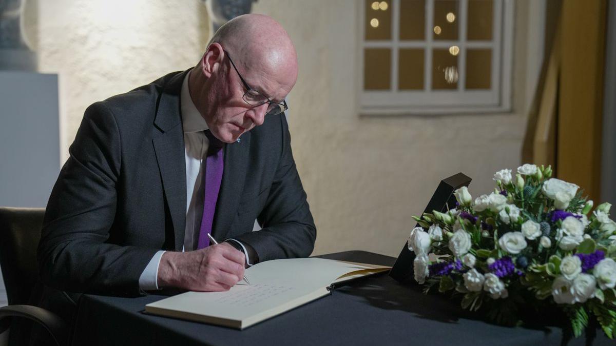 John  Swinney, in a dark suit and purple tie, signs the condolence book which is on a table with a black cloth and bouquet of flowers.