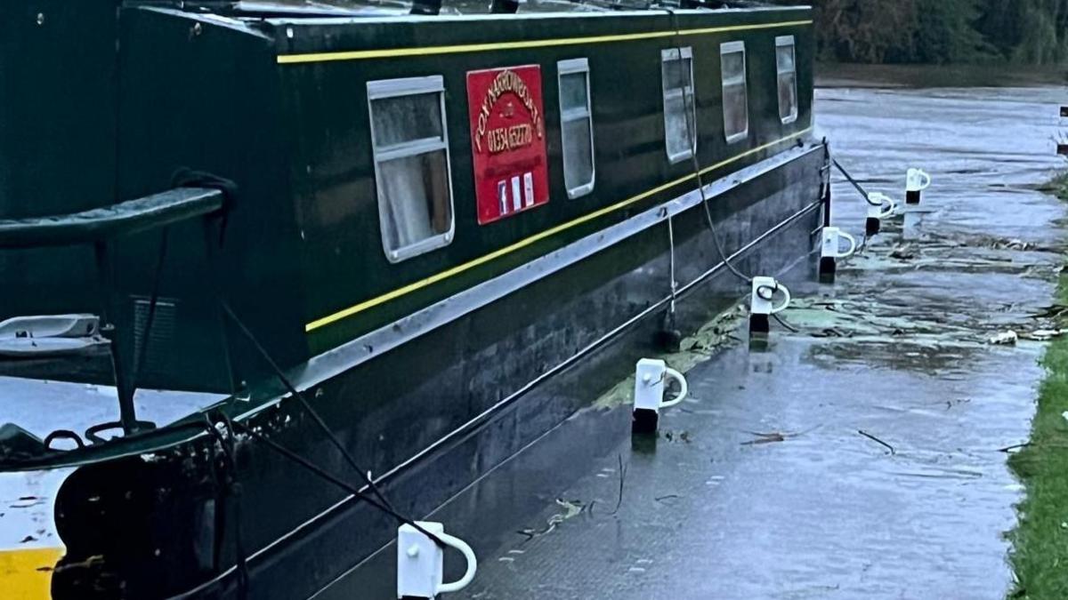 A 55-foot long black narrowboat moored to some posts on the river edge, the river has flooded over on to the pathway and weeds can be seen amongst the water there. 