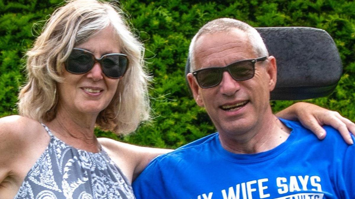 A woman with shoulder-length blonde hair, wearing hoop earrings, sunglasses and a paisley patterned dress stands with her arm around the shoulders of a man in a powerchair. He has short grey hair and is wearing a blue t-shirt and sunglasses.