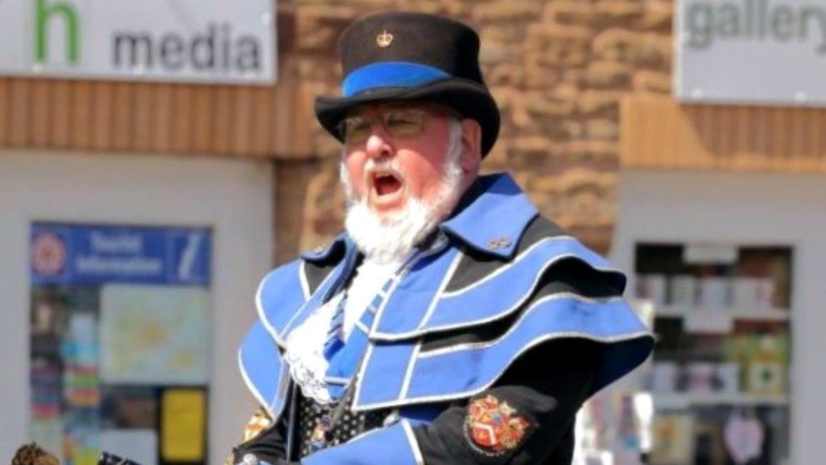 A man with a white beard is wearing a black top hat with a blue ribbon wrapped around the base. He is wearing a frilly black and blue jacket.