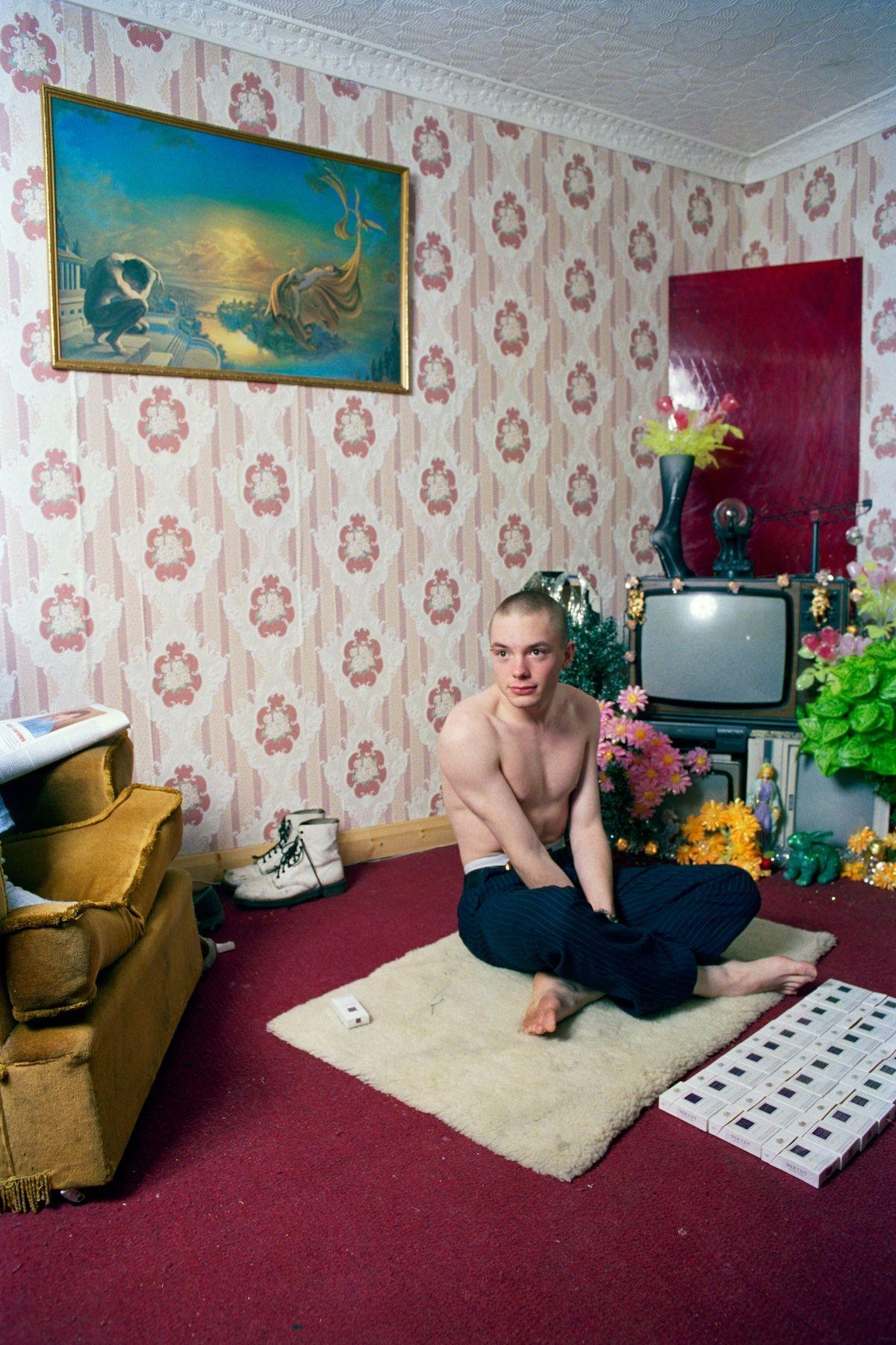 A photograph from the exhibition shows a young man, with cropped hair and no shirt sitting on the floor in a 90s living room