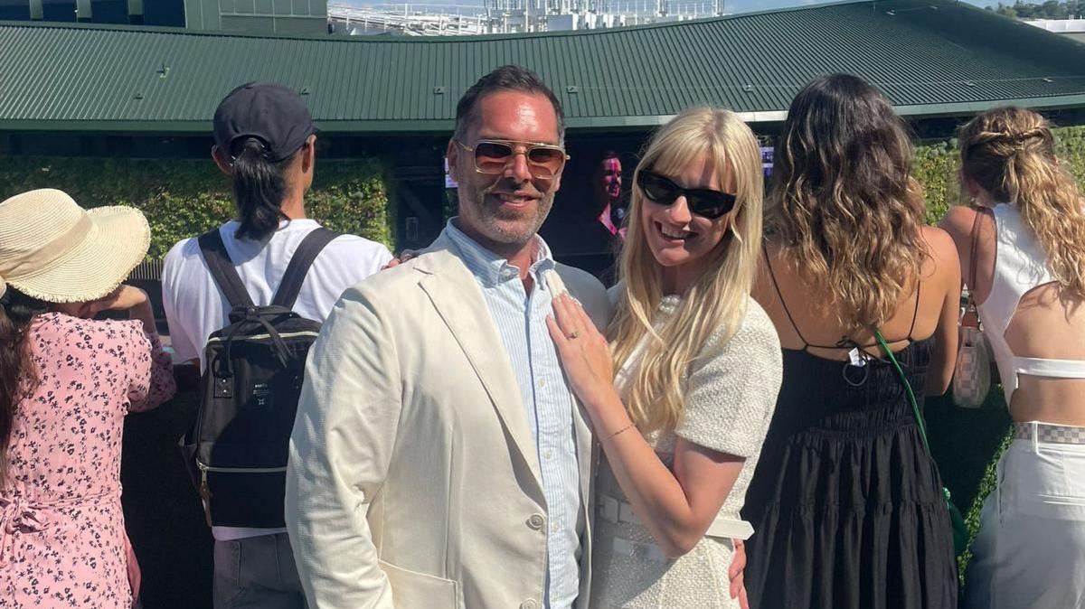 The couple wearing cream and white outfits and sunglasses, stood posing for a photograph as Ms Adams is resting her hand on Mr Barsdell's chest to display the ring. There are crowds behind them watching the tennis.