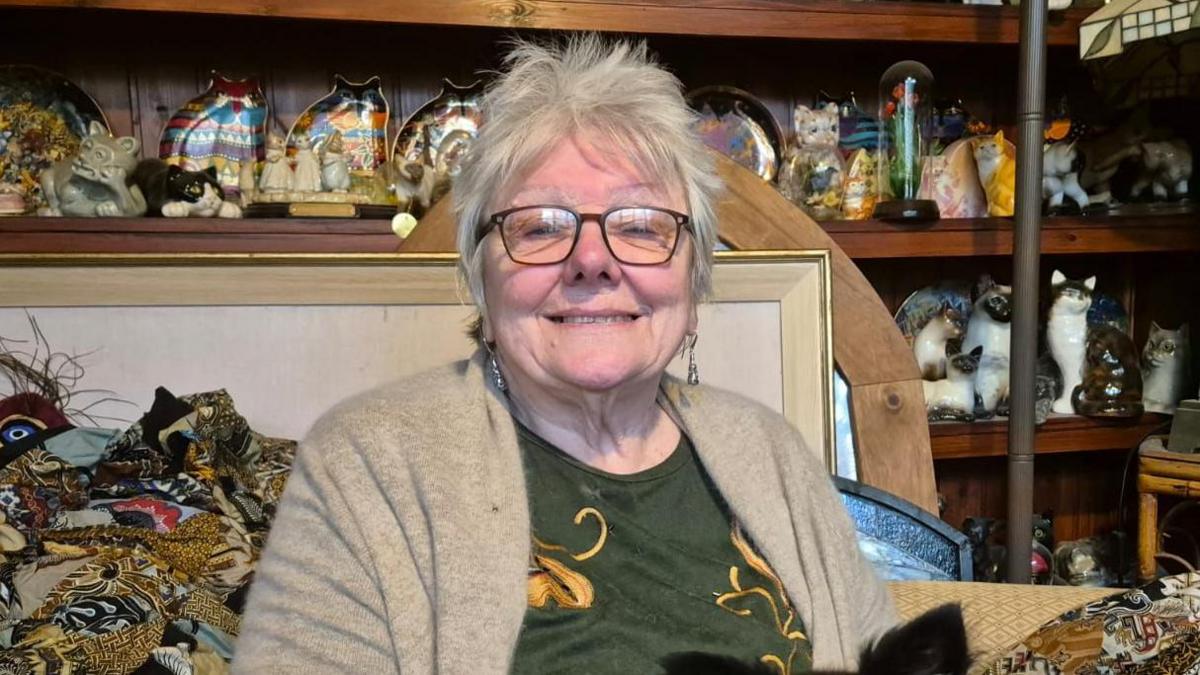 Jayne smiles into the camera. She is sat in her house surrounded by the items she collects, including ornamental cats and pictures. She has glasses, short grey hair and is wearing a light brown cardigan over a green patterned top.